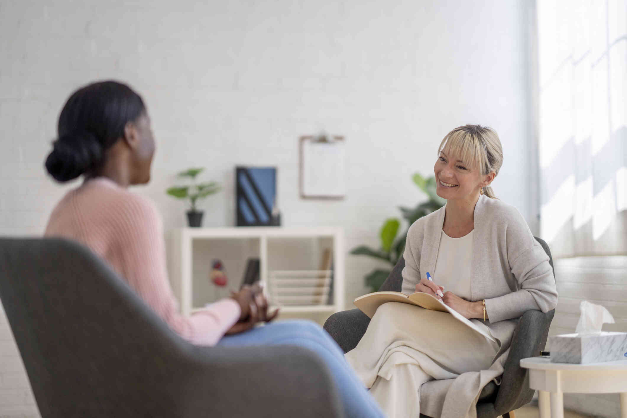 A woman in a pick shirt sits in a chair across from her female therapist who is smiling at her.